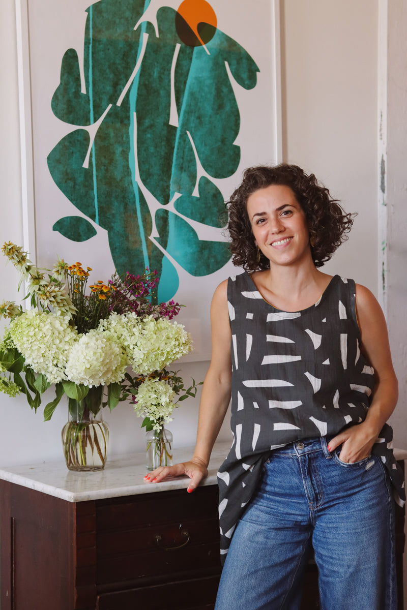 Leila Simon Hayes standing in her studio with her large wall art piece and floral arrangement.