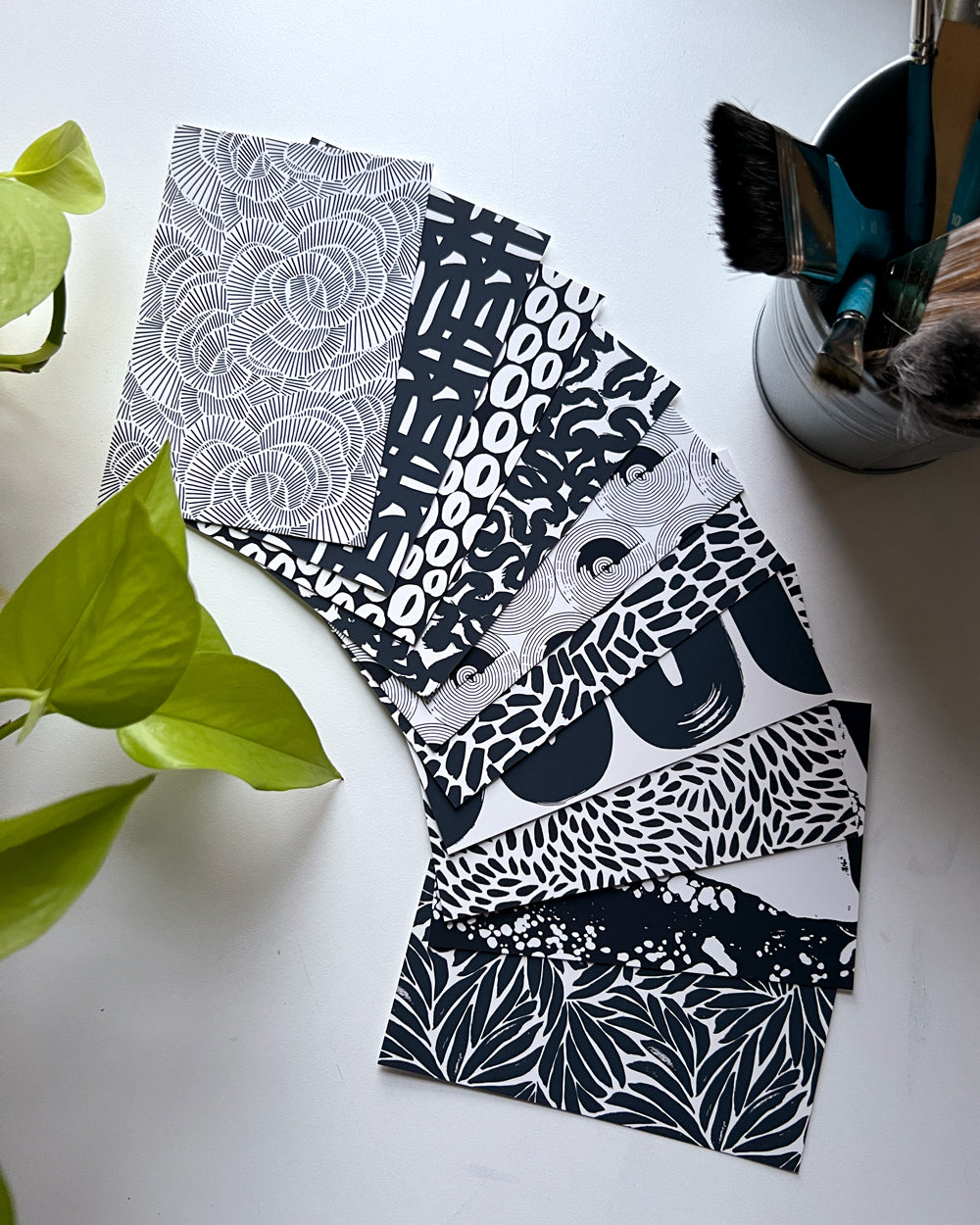 A stack of 10 postcards with various black and white floral and geometric patterns, displayed on a table with a plant in the background.