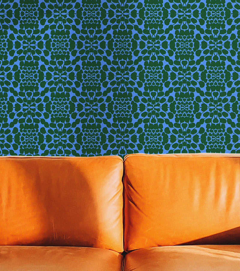 A close-up of an orange leather sofa seat and back cushions against a blue and green patterned wallpaper background. The wallpaper design is botanical and floral, designed by artist Leila Simon Hayes. 
