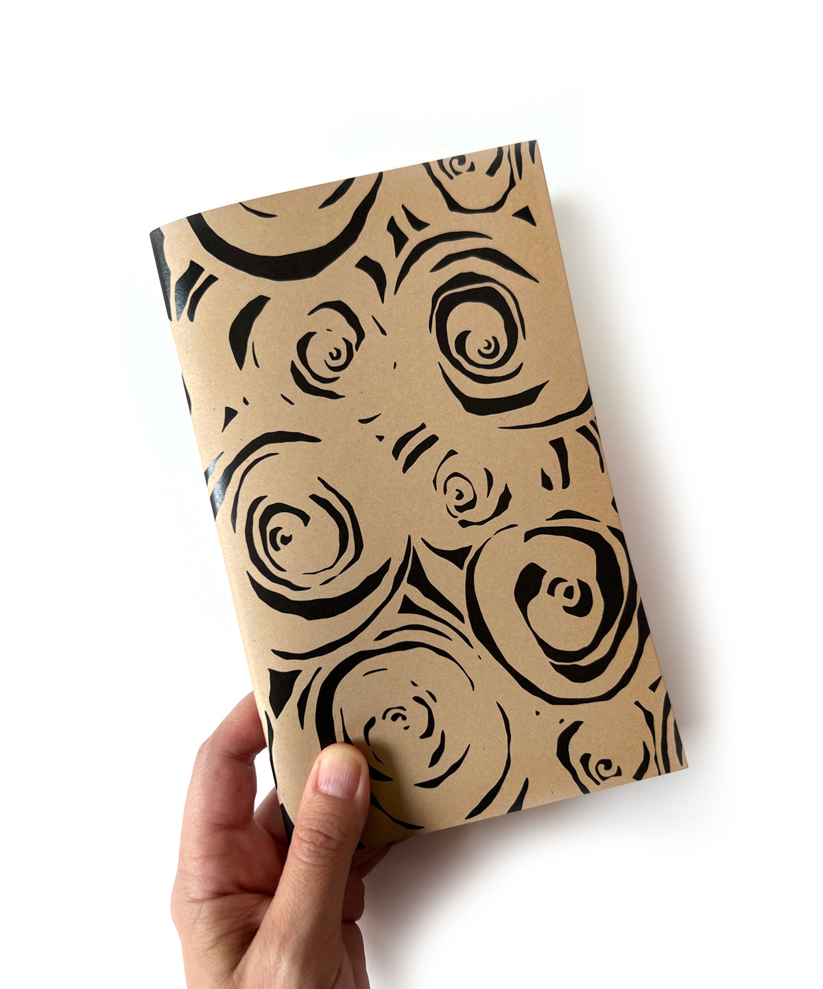 Notebook with black abstract rose pattern on tan cover, held by a hand on a white background. Designed by artist Leila Simon Hayes.
