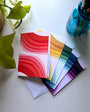 Five folded card packs with different colors of the rainbow. Matching envelopes are behind the cards spread out. Designed by artist Leila Simon Hayes.
