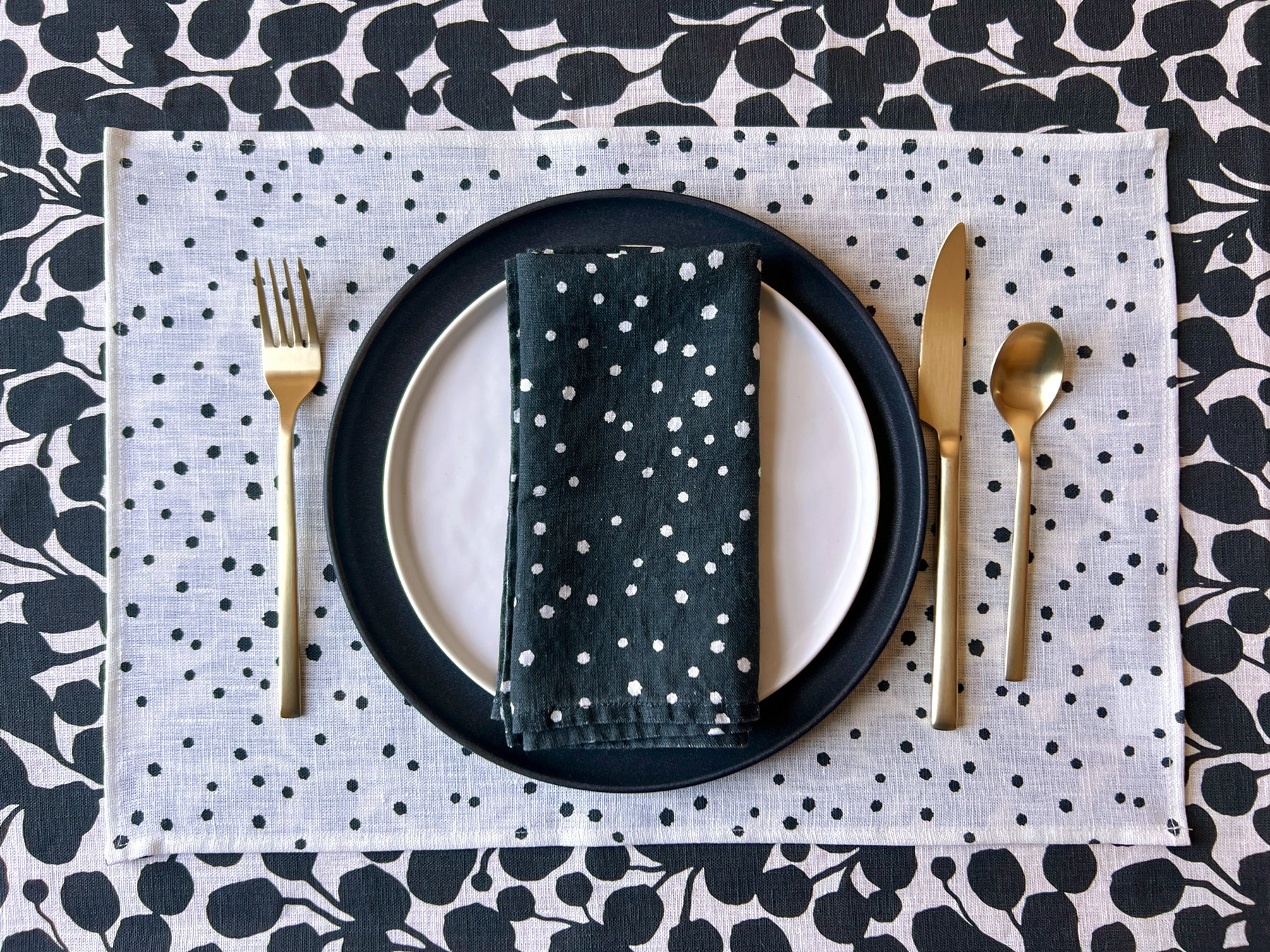 A white linen placemat with a dotted pattern, displayed on a table with a black plate, gold cutlery, and a black napkin with white organic dots folded in the center.All over a black and white botanical tablecloth. Table linens designed by artist Leila Simon Hayes