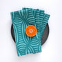Stack of teal striped patterned napkins designed by artist Leila Simon Hayes with a halved orange on a black plate against a white background