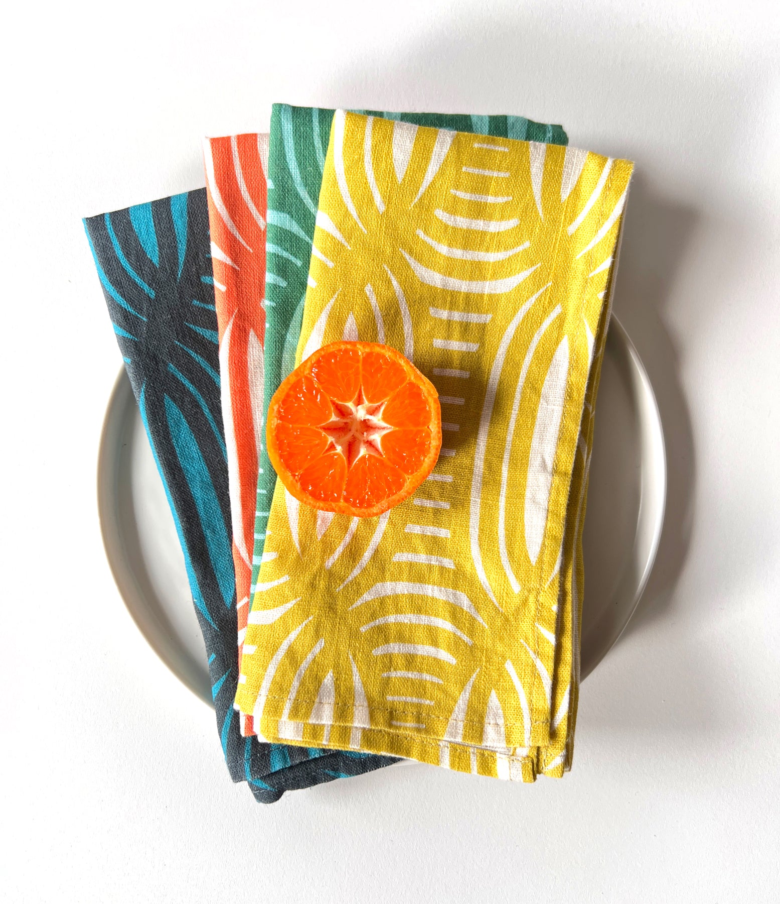 Four layered dinner napkins with a geometric pattern. Each napkin is a different color, yellow, green, blue and coral. Designed by artist Leila Simon Hayes