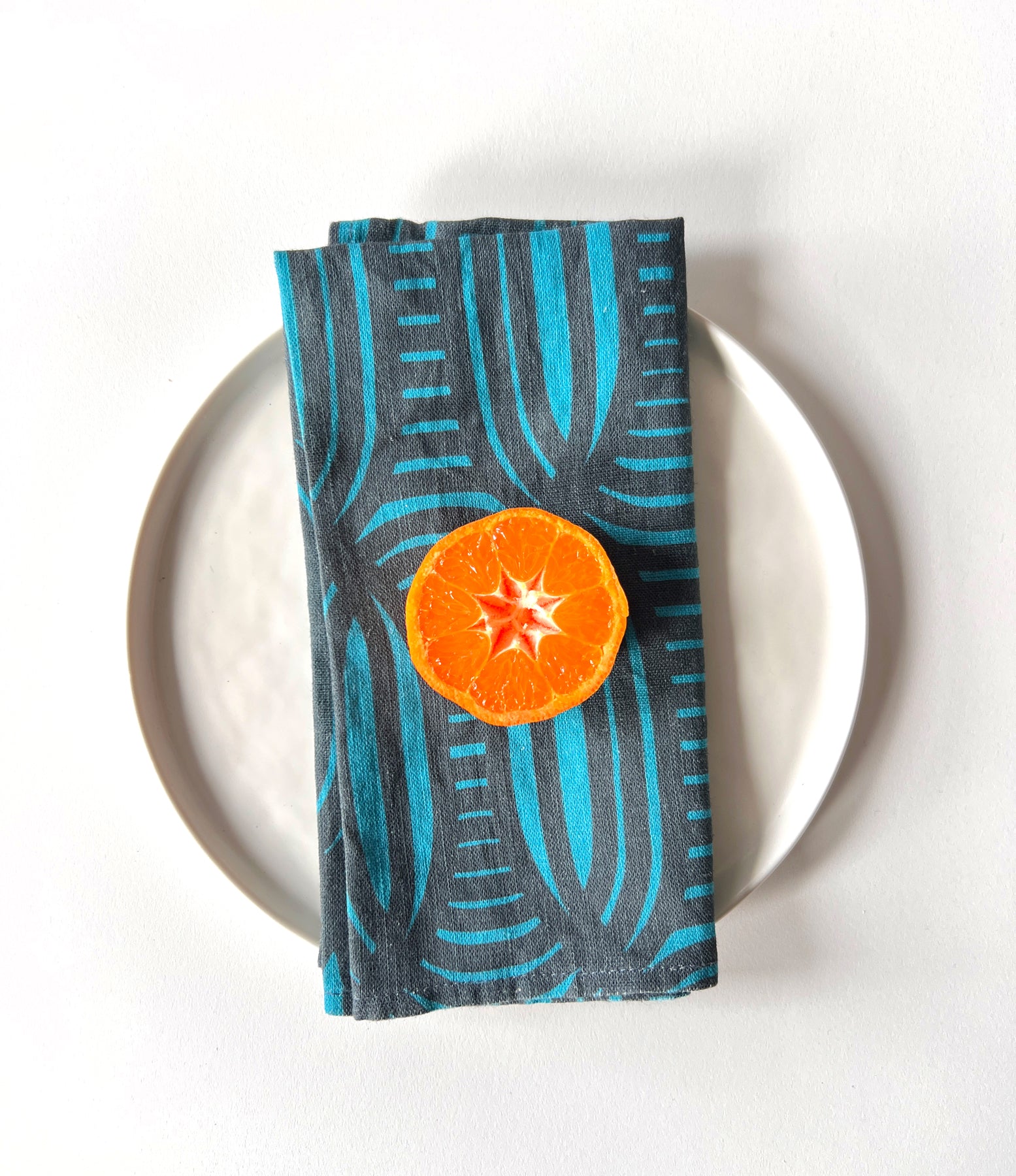 A blue linen napkin with a geometric pattern is displayed on a white plate, with a slice of orange placed on top of it. designed by artist Leila Simon Hayes