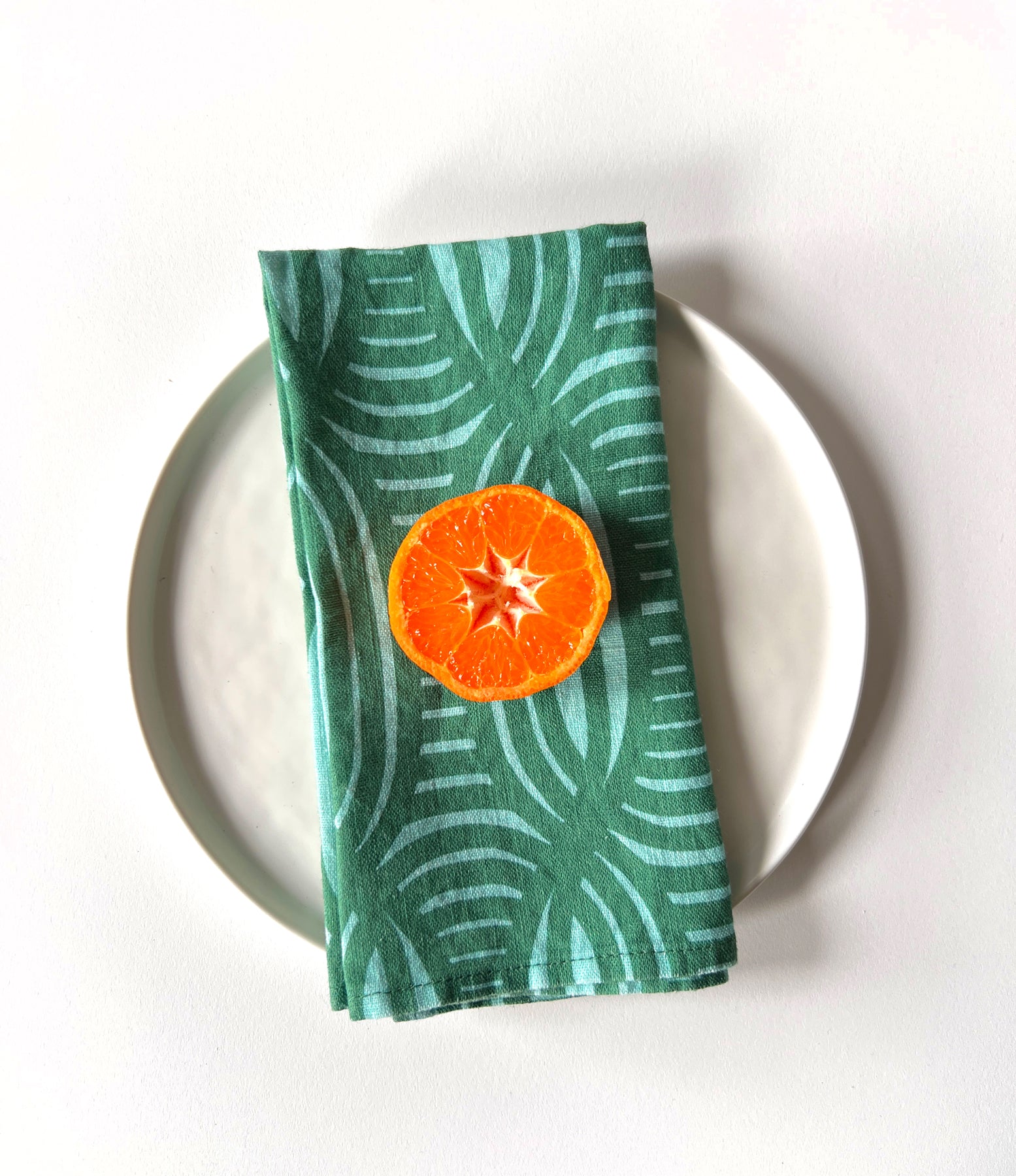 A green linen napkin with a striped geometric pattern, a slice of orange placed on top of it. Designed by artist Leila Simon Hayes