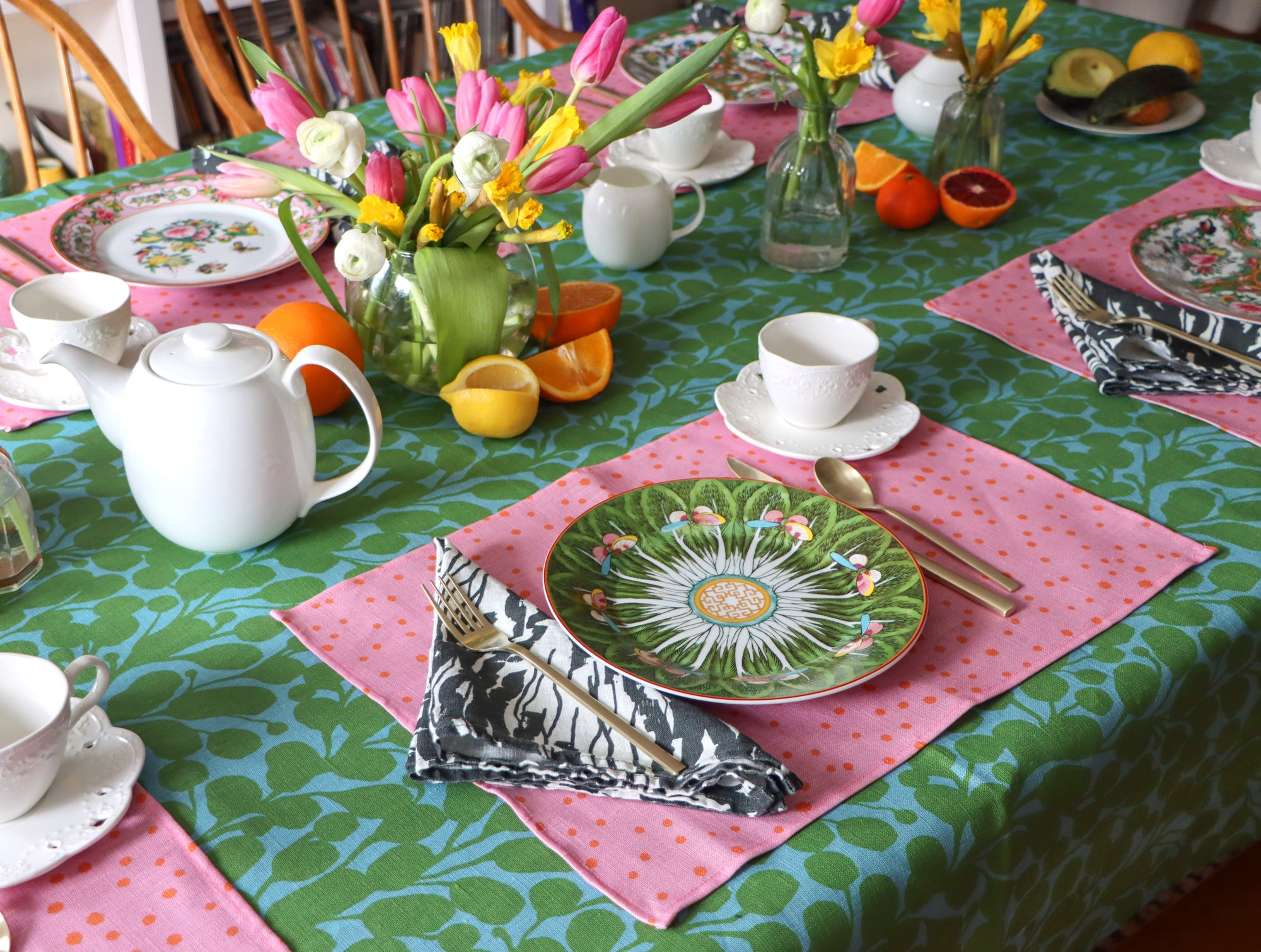 A set table with a black and white wave napkin design, a pink linen placemat, and the tablecloth is green and blue with a floral pattern. Designed by artist Leila Simon Hayes