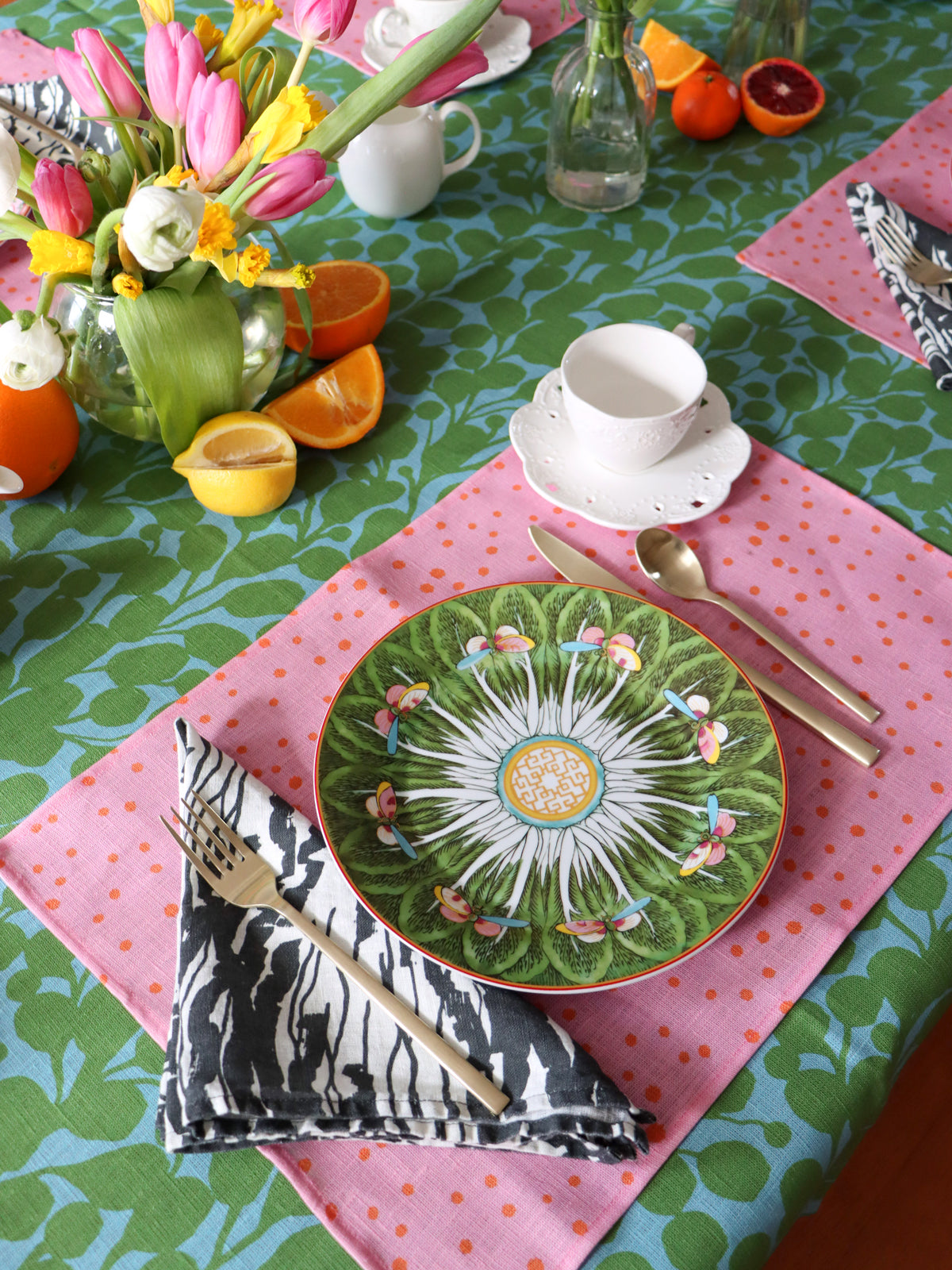 A set table with a black and white wave patterned napkin, pink polka dot placemat, and green and blue floral tablecloth. Designed by artist Leila Simon Hayes.