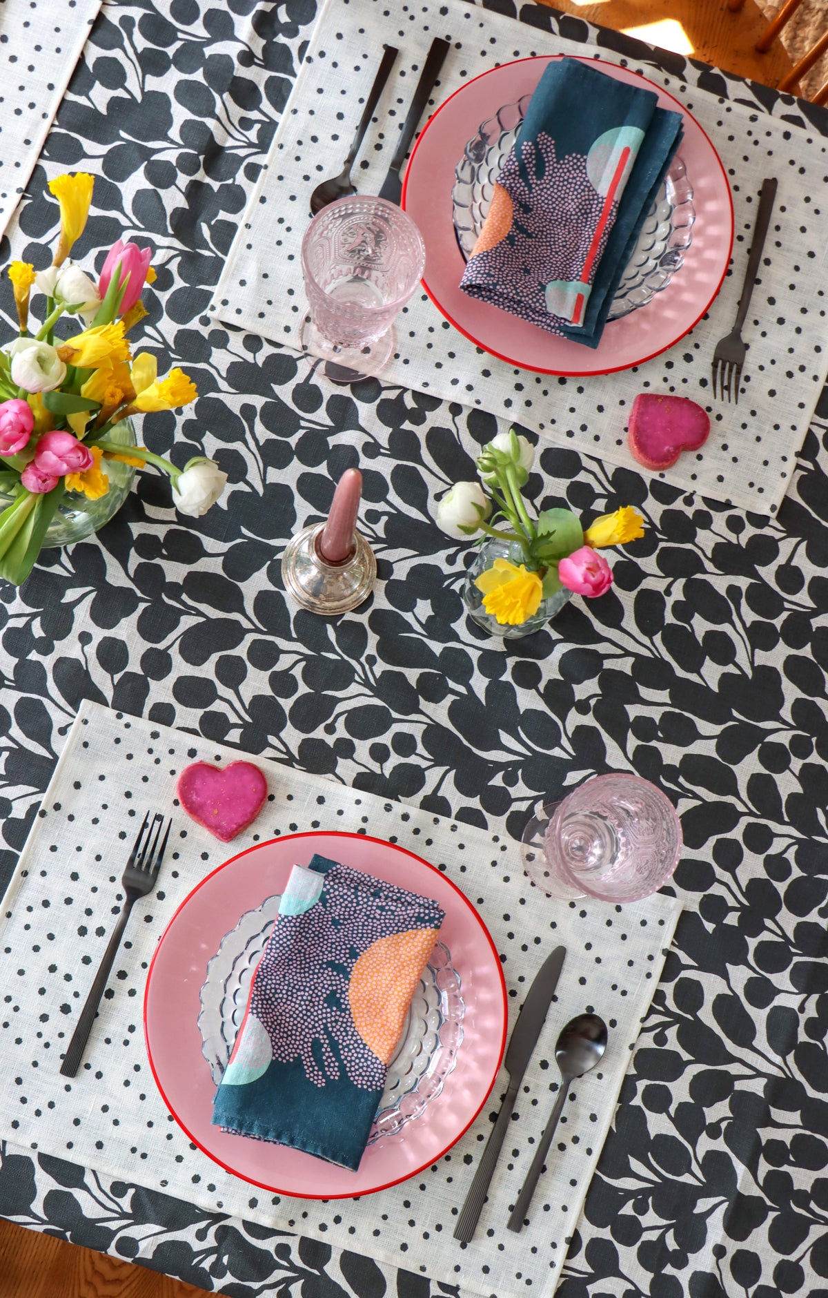 A black and white floral linen tablecloth with polka dot placemats and blue napkins, designed by artist Leila Simon Hayes. Flowers, candles, plates, and cutlery are also set around the table. 