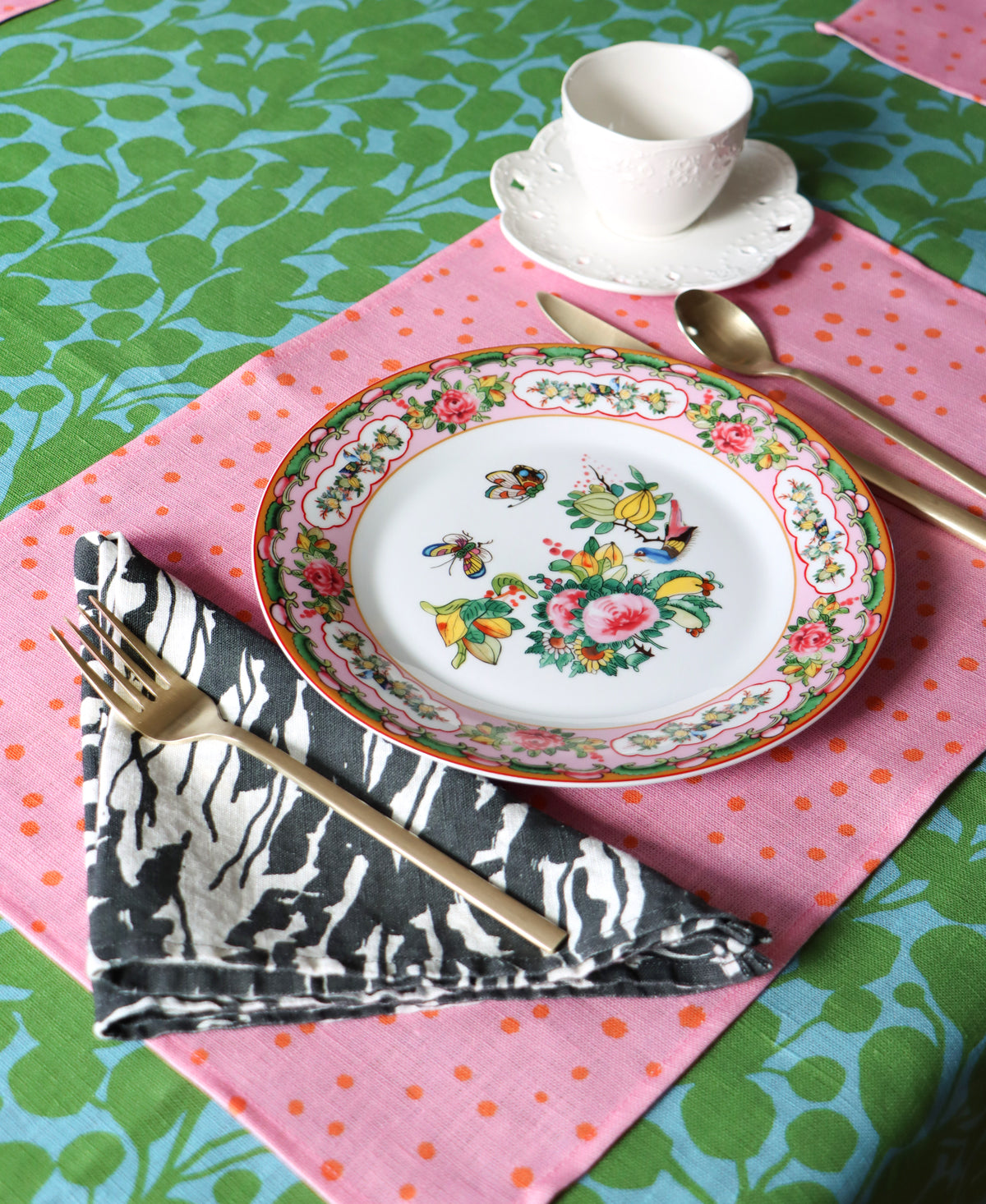 A set table with a black and white wave napkin design, a pink linen placemat, and the tablecloth is green and blue with a floral pattern. Designed by artist Leila Simon Hayes