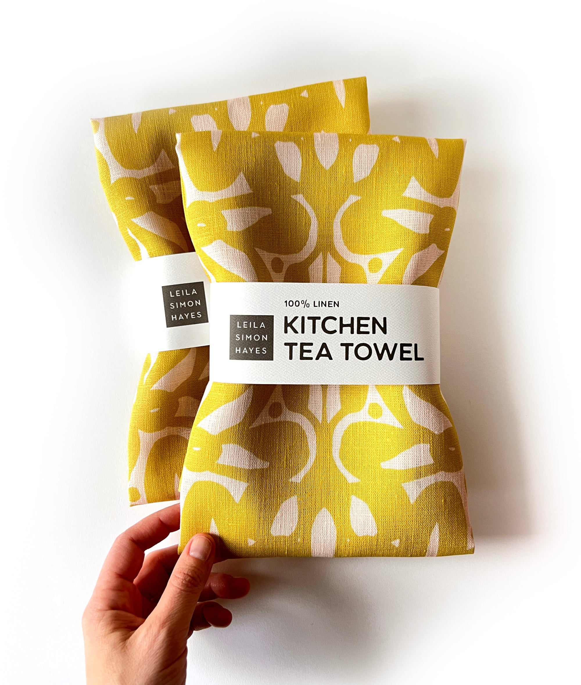 Yellow abstract, patterned, kitchen tea towels with labels saying "100% linen Kitchen Tea Towels" on a white background. Designed by artist Leila Simon Hayes.