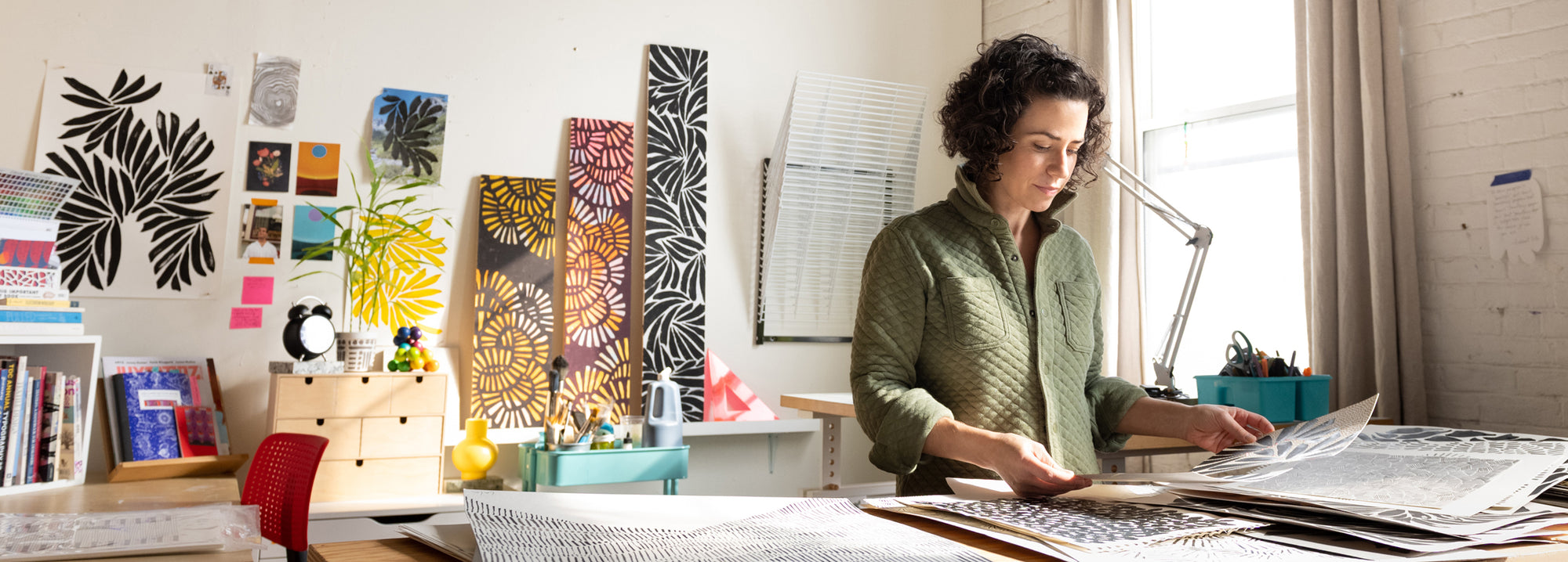 Leila Simon Hayes in a green shirt stands at a table in a bright, creative studio, surrounded by art prints, books, and papers. This is where she designs her home decor.